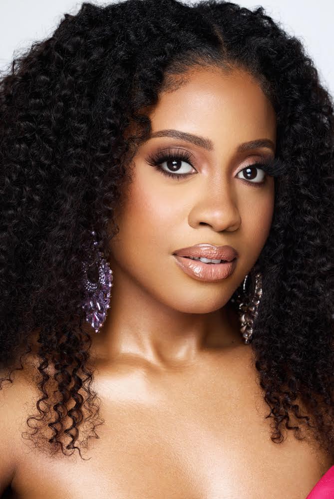 A portrait of Ludnie Augustin showcasing her natural curly hair and bold makeup, with a focus on her expressive eyes and glossy lips, complemented by elegant earrings.